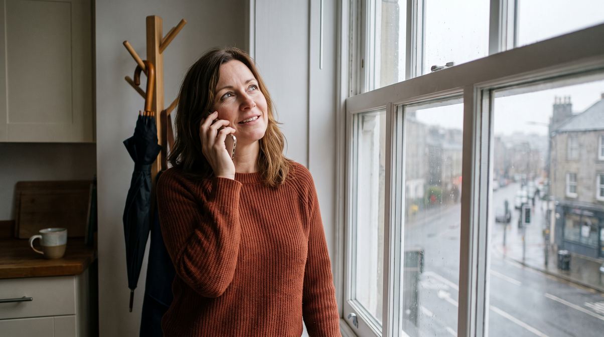 A woman on the phone at her kitchen window on a rainy morning, smiling up at the sky
